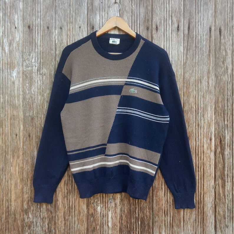 May include: A blue and brown striped sweater with a green crocodile logo. The sweater has a crew neck and long sleeves.