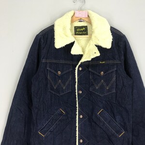 Op de afbeelding: Een donkerblauw denim jack met een sherpa voering. Het jack heeft een knoopsluiting, twee borstzakken en twee voorzakken. Het label op het jack staat "Wrangler" en "Wrangler Chaps".