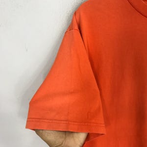 Pu&ograve; includere: Primo piano del tessuto arancione di una t-shirt. Il tessuto &egrave; liscio e ha una leggera lucentezza.