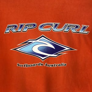 Pu&ograve; includere: Una maglietta rossa con le parole "RIP CURL" in lettere blu e bianche. Sotto il testo c'&egrave; un logo d'onda blu e bianco all'interno di un rombo. Le parole "Surfboards Australia" sono stampate sotto il logo.