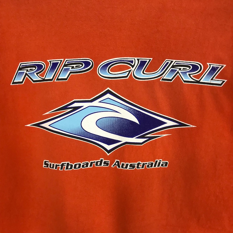 Pu&ograve; includere: Una maglietta rossa con le parole "RIP CURL" in lettere blu e bianche. Sotto il testo c'&egrave; un logo d'onda blu e bianco all'interno di un rombo. Le parole "Surfboards Australia" sono stampate sotto il logo.