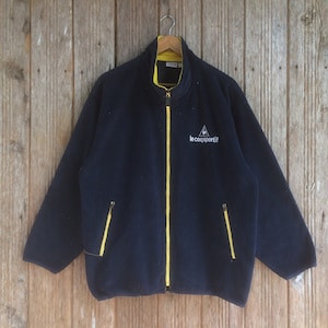 Op de afbeelding: Een marineblauwe fleecejack met een gele rits en een wit logo met de tekst "le coq sportif" op de voorkant.