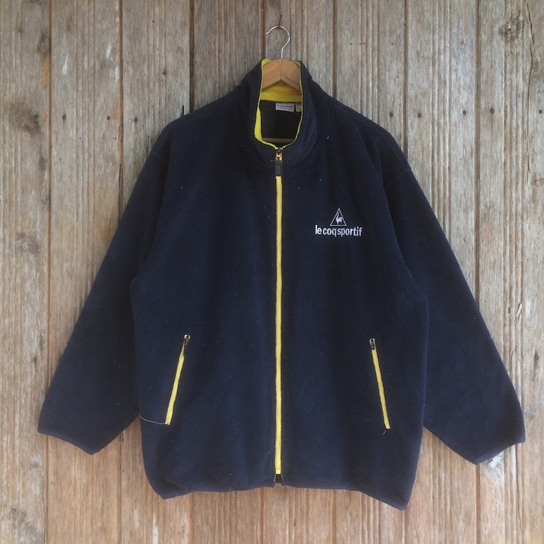 Op de afbeelding: Een marineblauwe fleecejack met een gele rits en een wit logo met de tekst "le coq sportif" op de voorkant.