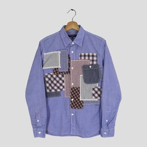 May include: A light blue long-sleeved shirt with a patchwork design. Various fabric patches in different patterns, such as stripes and checks, are sewn onto the front. The shirt has a button-down collar and cuffs.
