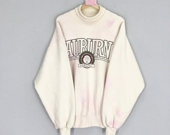May include: Cream-colored sweatshirt with the word "AUBURN" in large, black block letters across the chest. Below the word is a circular emblem with text and the words "SINCE 1856". The sweatshirt has a mock neck and long sleeves.
