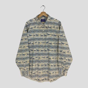 May include: A long-sleeve button-down shirt with a repeating horse pattern in shades of blue, tan, and cream. The shirt has a collared neckline and a single chest pocket. The fabric appears to be a woven material.