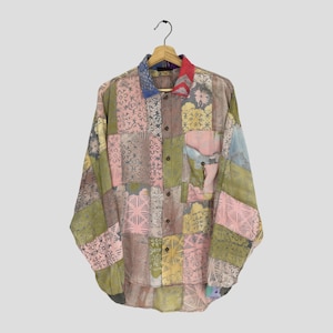 May include: A long-sleeved patchwork shirt in pastel shades of pink, green, and yellow. The shirt has a button-down front, a chest pocket, and a collar with blue and red details. The shirt is displayed on a wooden hanger.