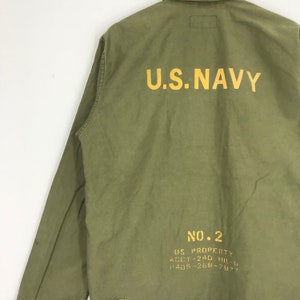 May include: Olive green military-style jacket with the text "U.S. NAVY" printed in yellow across the back. Additional yellow text includes "NO. 2" and other details. The jacket has long sleeves and a collar.