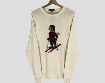 May include: A cream-colored knitted sweater with a brown bear skiing on the front. The bear is wearing a red and black plaid scarf. The sweater has a crew neck and long sleeves.
