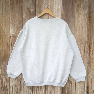 May include: A grey sweatshirt with long sleeves and a crew neck. The sweatshirt is hanging on a wooden hanger.