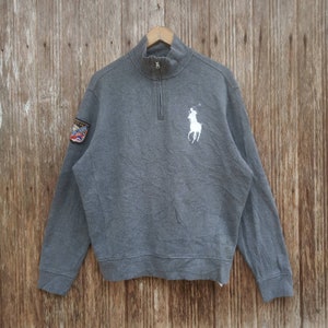 May include: A grey long-sleeved sweatshirt with a white polo player logo on the chest and a small patch on the left sleeve. The sweatshirt has a quarter-zip closure.