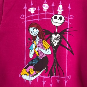 Puede incluir: Una camiseta rosa con un estampado gr&aacute;fico de Jack Skellington y Sally de Pesadilla antes de Navidad. Los personajes est&aacute;n de pie frente a una puerta negra con un dise&ntilde;o de coraz&oacute;n.