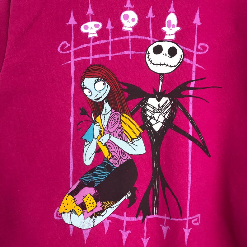 Puede incluir: Una camiseta rosa con un estampado gr&aacute;fico de Jack Skellington y Sally de Pesadilla antes de Navidad. Los personajes est&aacute;n de pie frente a una puerta negra con un dise&ntilde;o de coraz&oacute;n.