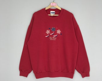 May include: Red sweatshirt with a white, blue, and red embroidered design of stars and ribbons. The text "God Bless America" is embroidered below the design.