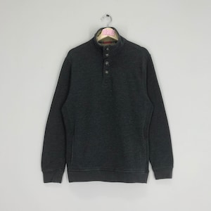 Puede incluir: Una sudadera gris oscuro con cuello abotonado. La sudadera tiene un forro polar y tres botones en la parte delantera. La prenda está colgada de una percha de metal sobre un fondo gris claro. La sudadera tiene mangas largas y un dobladillo acanalado.