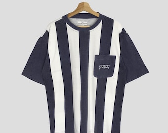Könnte beinhalten: Ein marineblaues und weiß gestreiftes T-Shirt mit einer kleinen Tasche auf der linken Seite. Die Tasche ist mit dem Wort "GUESS" bestickt.