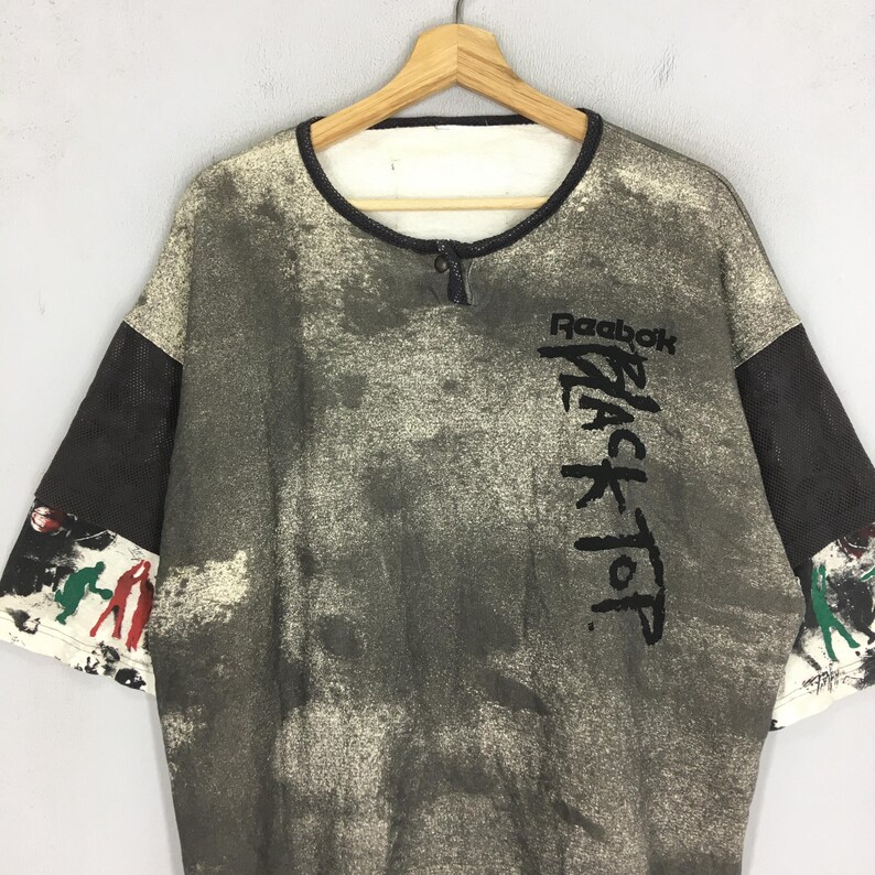May include: A vintage Reebok Black Top t-shirt with a distinctive, distressed pattern. The shirt has a black collar, sleeves, and the Reebok logo with "Black Top" printed vertically. The sleeves have a mesh design and colourful graphics.