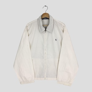 Puede incluir: Chaqueta blanca con cremallera, cuello y mangas largas. Un pequeño logotipo azul marino está en el pecho izquierdo. La chaqueta está colgada de una percha de madera.