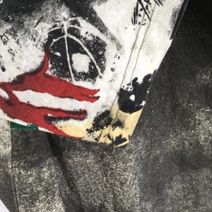 May include: Close-up of a section of a garment with a white, black, and red abstract design. The fabric has a distressed, speckled appearance. The edge of the garment is visible, showing stitching and a contrasting colour.