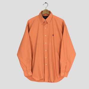 Op de afbeelding: Een lange mouwen, oranje overhemd met knoopsluiting. Het overhemd heeft een button-down kraag, een klein geborduurd logo op de borst en manchetten met knopen. Het overhemd hangt aan een houten hanger.