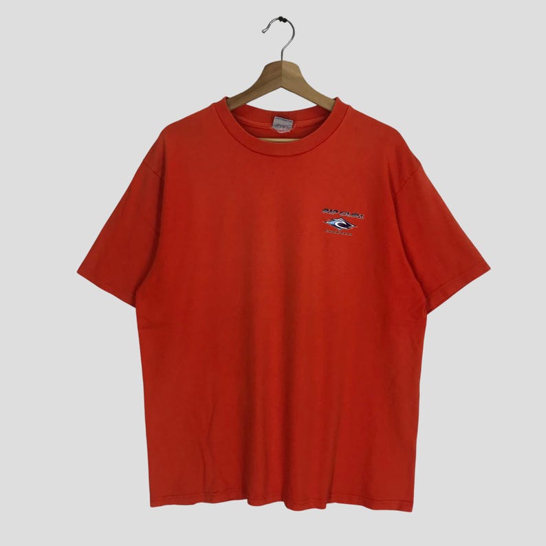 Pu&ograve; includere: Maglietta arancione a maniche corte con un piccolo logo ricamato sul davanti. Il logo &egrave; un design blu e bianco con il testo "Rip Curl" e "Australia".