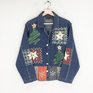 Może przedstawiać: Niebieska kurtka dżinsowa z patchworkowym wzorem przedstawiającym choinki, gwiazdy i kratę. Kurtka zapinana jest na guziki i ma długie rękawy.