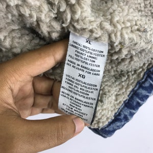 Puede incluir: Una chaqueta vaquera azul con forro de sherpa. La etiqueta dice "XL" y enumera el contenido de la tela como 100% algod&oacute;n para la c&aacute;scara, 100% poli&eacute;ster para el forro, 100% algod&oacute;n para el forro del bolsillo y 100% poli&eacute;ster para el relleno. La etiqueta tambi&eacute;n dice "Hecho en Bangladesh" y "Ver el reverso para el cuidado".