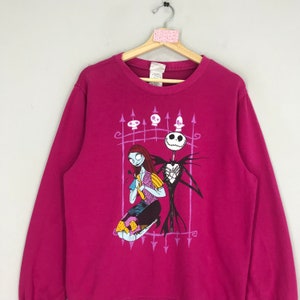 Puede incluir: Una sudadera rosa con un estampado gr&aacute;fico de Jack Skellington y Sally de Pesadilla antes de Navidad. El estampado presenta una valla morada con calaveras y flechas.