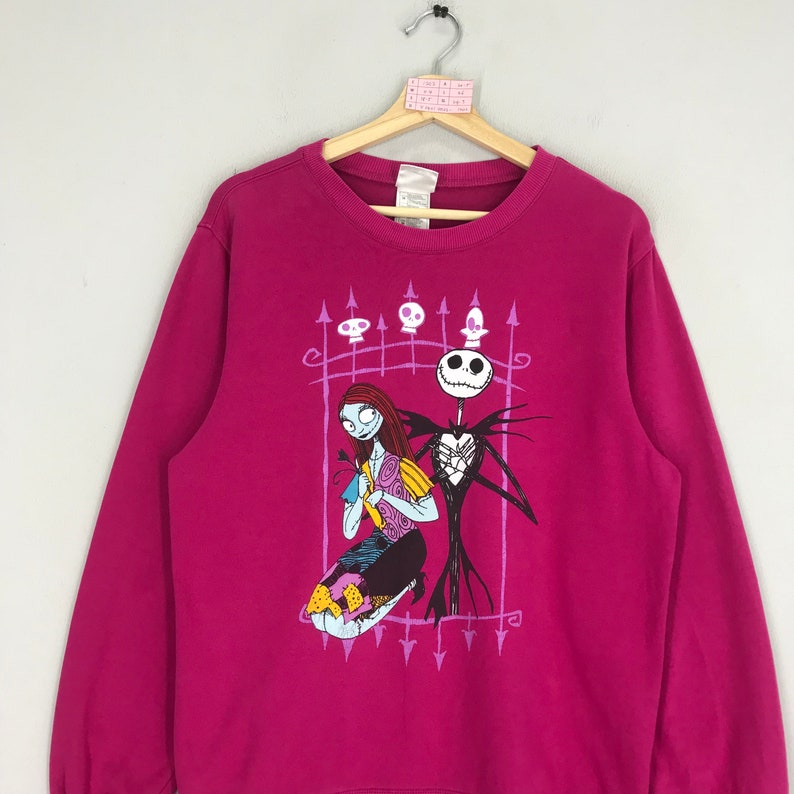 Puede incluir: Una sudadera rosa con un estampado gr&aacute;fico de Jack Skellington y Sally de Pesadilla antes de Navidad. El estampado presenta una valla morada con calaveras y flechas.