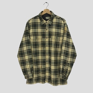 Puede incluir: Una camisa de franela a cuadros de manga larga en un esquema de color amarillo y verde. La camisa tiene un frente abotonado y dos bolsillos en el pecho.