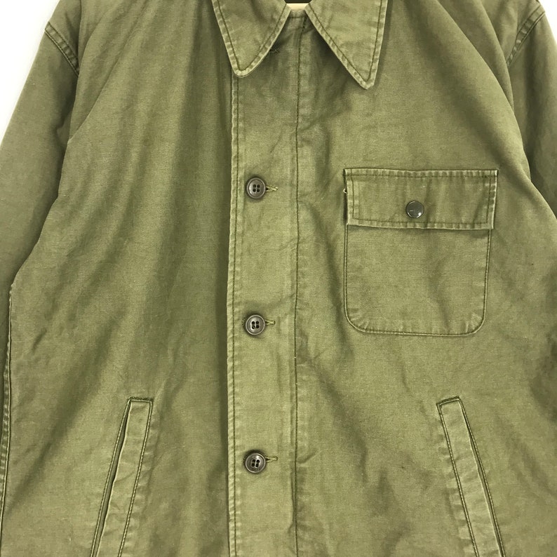 May include: Olive green jacket with a button-up front, a collared neckline, and a single chest pocket. The jacket has two side pockets and a relaxed fit. The fabric appears to be a durable, possibly canvas material.