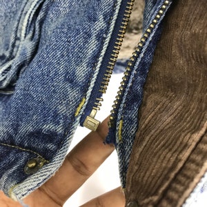 Puede incluir: Primer plano de una cremallera de una chaqueta vaquera con forro de pana marr&oacute;n. La cremallera es de metal y tiene un acabado dorado.