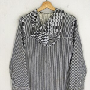 May include: A blue and white striped hooded jacket hanging on a wooden hanger. The jacket has long sleeves and a front pocket. The fabric has vertical stripes. The jacket is made of a lightweight material.