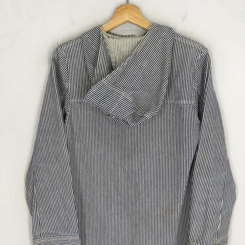 May include: A blue and white striped hooded jacket hanging on a wooden hanger. The jacket has long sleeves and a front pocket. The fabric has vertical stripes. The jacket is made of a lightweight material.