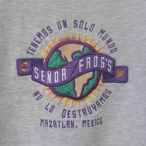 May include: A grey sweatshirt with a colourful embroidered logo. The logo features a globe with a purple banner that reads "Se&ntilde;or Frog's". The text around the globe reads "Tenemos un solo mundo no lo destruya mos Mazatlan, Mexico".