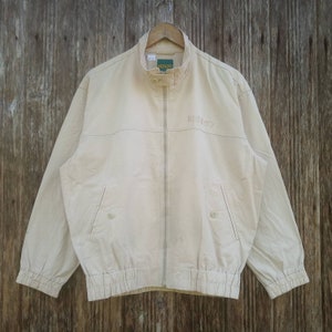 May include: A beige zip-up jacket with a ribbed waistband and cuffs. The jacket has a green label with the text "KENZO" and the text "KENZO" embroidered on the front in a light pink colour.