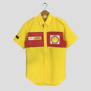 以下が含まれることがあります： 赤のアクセントが付いた黄色の半袖シャツ。シェルロゴパッチと「Formula Shell」パッチが付いています。左袖には小さな黒いパッチが付いています。シャツは木製のハンガーに吊るされています。