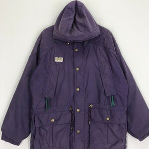 May include: A purple hooded jacket with a front zipper closure and multiple pockets. The jacket features a drawstring at the waist and a small logo patch on the chest. The jacket has gold-colored buttons and green accents.