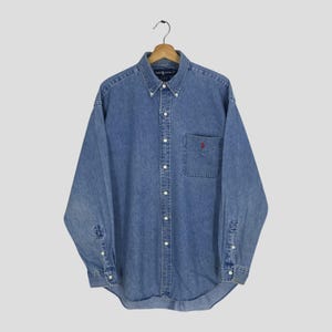 Puede incluir: Una taza de cerámica blanca con un asa negra y el texto "But First, Coffee" en un estilo manuscrito. La taza está sobre una superficie blanca. La taza mide aproximadamente 10 cm de alto.