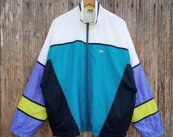 Pode incluir: Uma jaqueta corta-vento Nike vintage com um design de blocos de cores branco, preto, turquesa e roxo. A jaqueta tem um fecho de correr e um logotipo da Nike no peito.