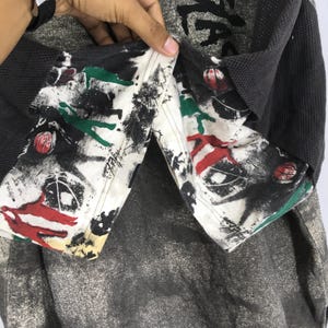 May include: A close-up of a grey and black long-sleeved shirt with a unique design. The shirt has a patterned collar with red, green, and black abstract art. The sleeves are a dark grey waffle knit, and the body is a mottled grey.