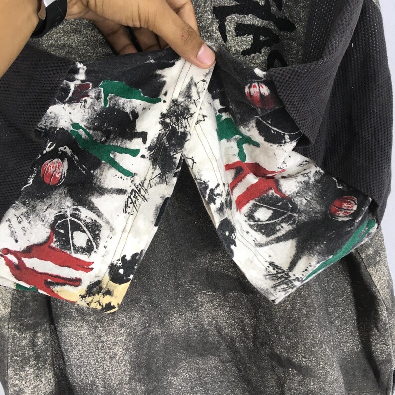 May include: A close-up of a grey and black long-sleeved shirt with a unique design. The shirt has a patterned collar with red, green, and black abstract art. The sleeves are a dark grey waffle knit, and the body is a mottled grey.