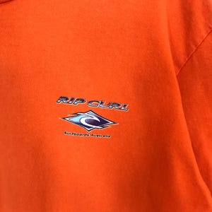 Pu&ograve; includere: Maglietta arancione con un logo Rip Curl blu e bianco e il testo "Surfboards Australia".