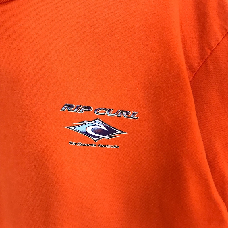 Pu&ograve; includere: Maglietta arancione con un logo Rip Curl blu e bianco e il testo "Surfboards Australia".