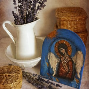 May include: A hand-painted wooden icon depicting an angel with wings, wearing a brown robe and a beaded necklace. The icon is set against a blue background with a gold halo and sunburst. The icon is displayed on a table with dried lavender and wicker baskets.