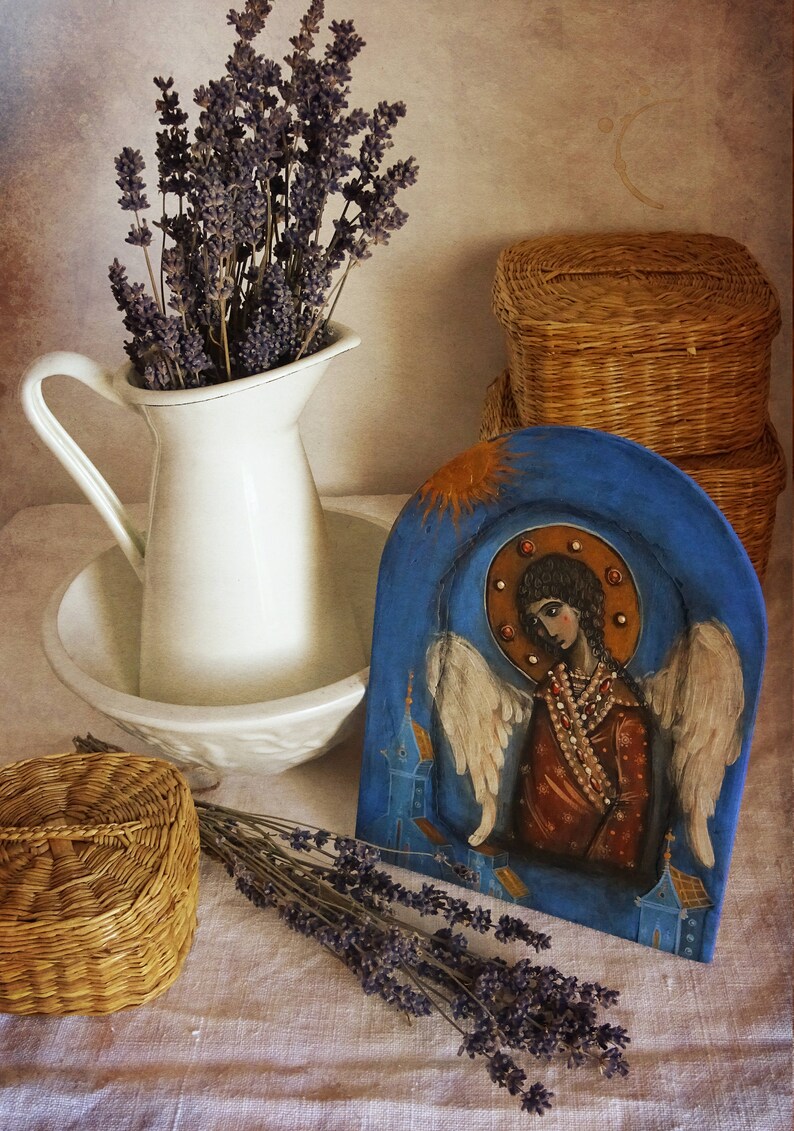 May include: A hand-painted wooden icon depicting an angel with wings, wearing a brown robe and a beaded necklace. The icon is set against a blue background with a gold halo and sunburst. The icon is displayed on a table with dried lavender and wicker baskets.