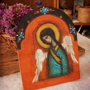 May include: A hand-painted wooden icon depicting an angel with wings, wearing a blue and gold robe. The angel has a halo and is set against a red background with a gold halo.