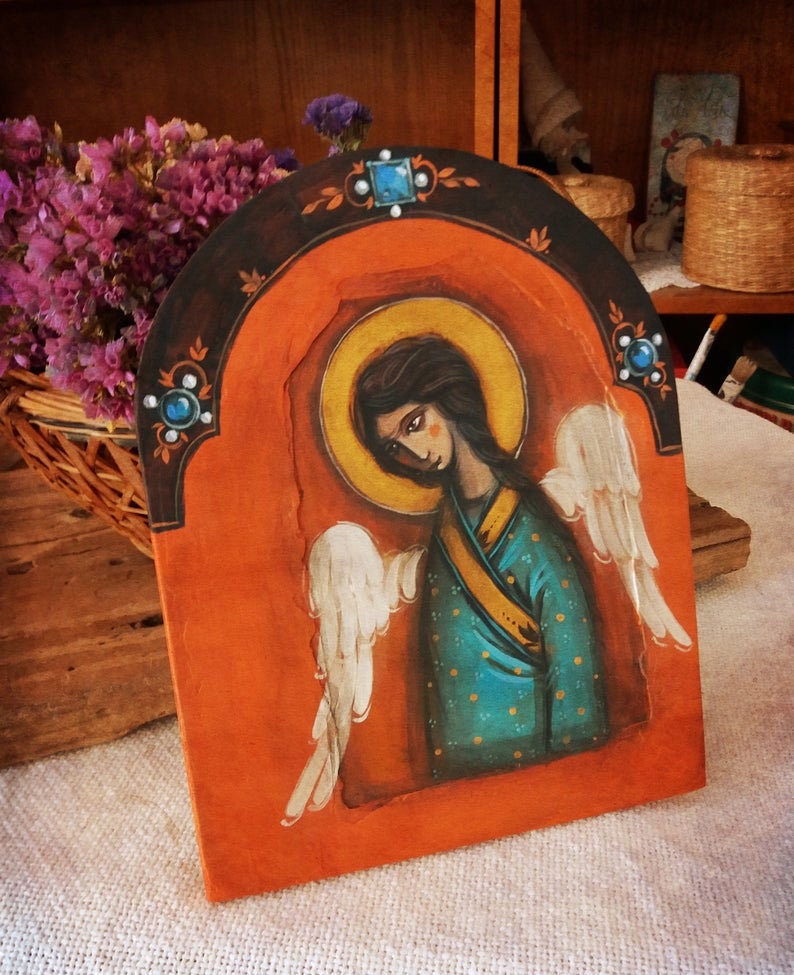 May include: A hand-painted wooden icon depicting an angel with wings, wearing a blue and gold robe. The angel has a halo and is set against a red background with a gold halo.