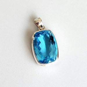 May include: A rectangular, light blue gemstone pendant with a silver-colored bail. The gemstone is a vibrant, clear blue, set in a polished silver frame. The pendant is displayed on a white surface.