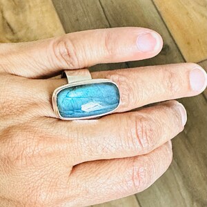 May include: A silver ring featuring a rectangular, iridescent blue gemstone. The gemstone displays a play of colors, with shades of blue and hints of white. The ring is set in a simple, modern silver band.